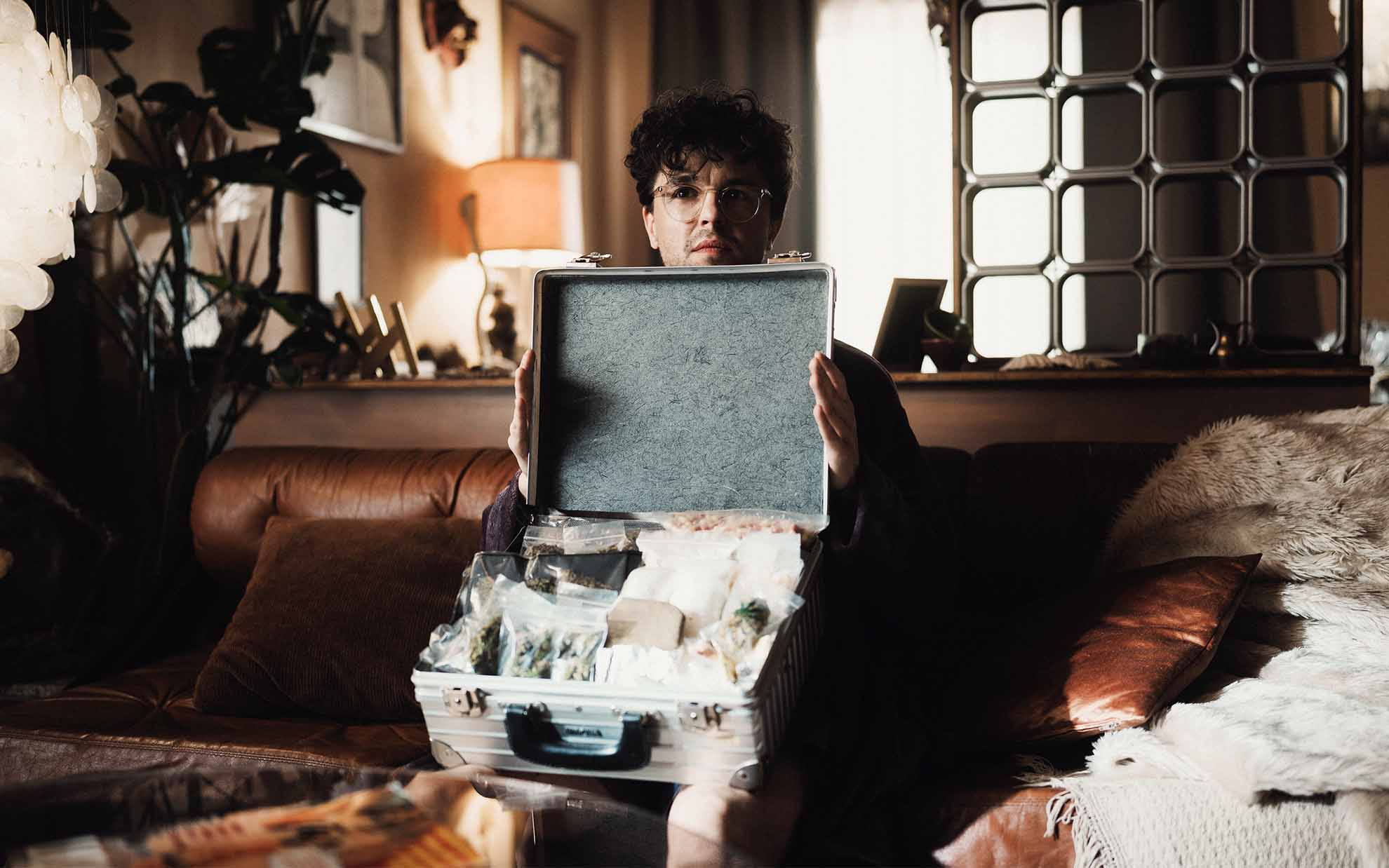 Picobello (2026) – Production Design for short film, main character on sofa presenting suitcase goods, warm domestic lighting, tense mood