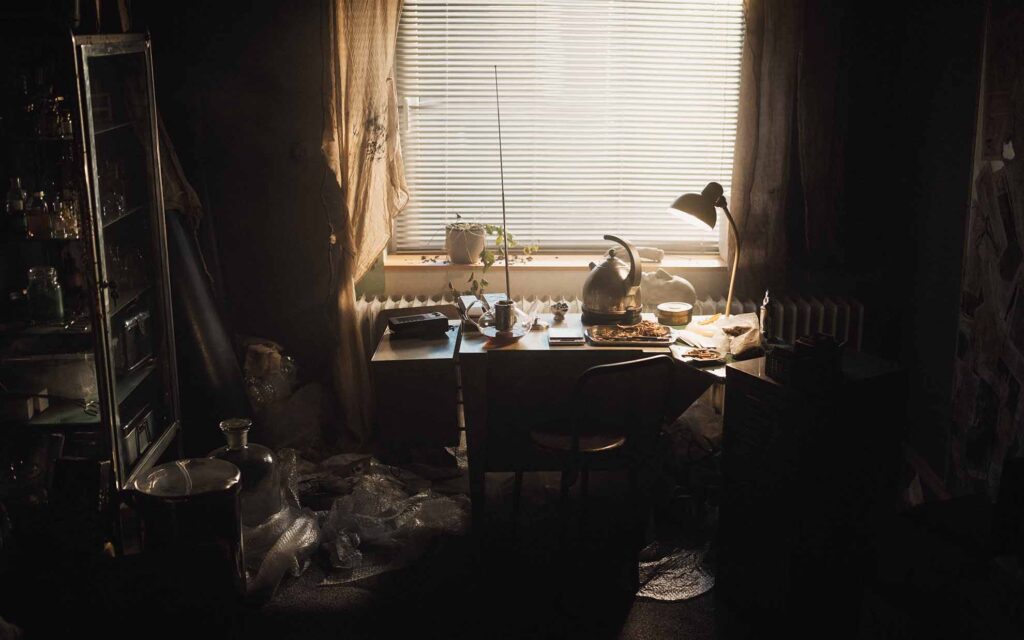 Picobello (2026) – Behind the Scenes Still - production design Picobello (2026) – Production Design for short film, Dim Improvised Lab with Warm Domestic Light, cluttered desk with retro phone, kettle and jars, plastic sheets, moody window light, tense yet homely space